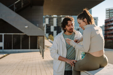 A man stands and talks while placing his fingers on the thigh of a woman who is sitting on the wall and clicking on the phone that shows
