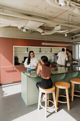 A female worker stands and holds a cup while talking to a female worker sitting across from her on a chair at a table while two male workers stand next to them
