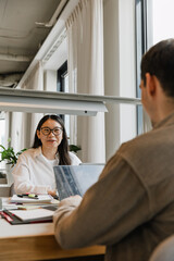 A female worker laughs and talks to a male worker across from her while they sit at a table in...