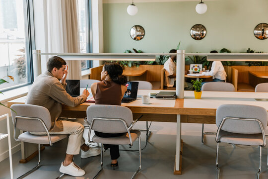 A male worker is talking to a female worker next to him while they are sitting on chairs at a table in front of laptops