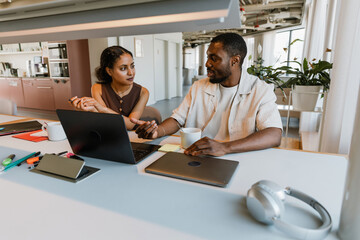 A male employee is talking to a female employee who is sitting next to him at the desk and holding...