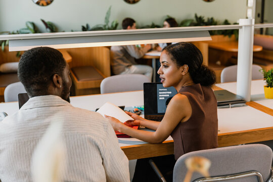 Female worker sitting on a chair and holding a notebook while talking to a male worker while they are sitting at a table