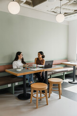 Two female workers are talking and sitting at a table while one of them is holding a notebook