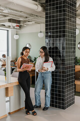 Two female workers stand and read a notebook that one of them is holding and talk while leaning on...