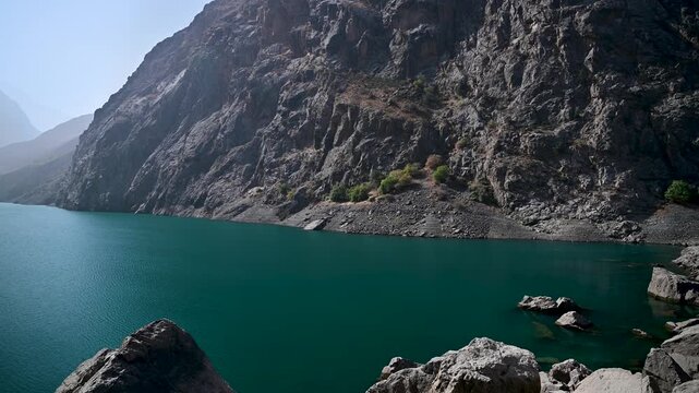 A pan left view of Nofin Lake, the fourth lake of the Seven Lakes in the Fann Mountains near Panjakent, Tajikistan, with steep rocky walls and deep blue mountain water under clear daylight.