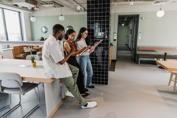 A male worker leans on a desk and stands next to two female workers who are reading a notebook and a folder