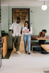 A male and female workers are reading a notebook she is holding as they walk past a female worker...