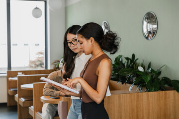A female worker is talking and pointing at a notebook which she is showing to a female worker...