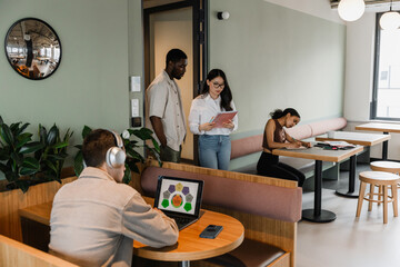 Male and female workers reading a notebook and walking past two workers sitting at desks while writing in a notebook and looking at a laptop