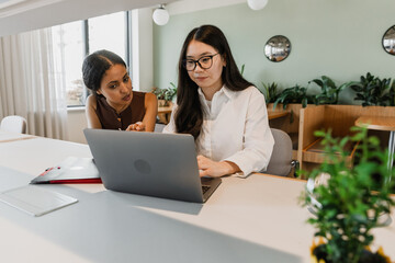 A female worker talks and points at a laptop that a female worker is typing on while they sit at a table