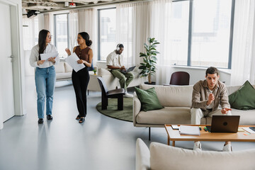 A male employee sits on a couch and looks at a laptop while two female employees walk by him carrying documents and talking