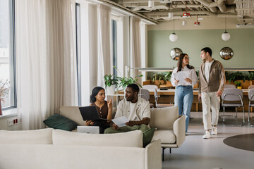 A female worker is holding a laptop and talking to a male worker who is listening to her and holding a document while they are sitting on a couch and two workers are walking towards them