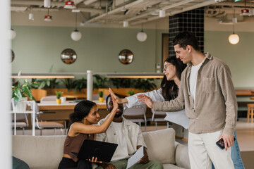 Male and female employees standing and giving high five to female employee holding laptop and sitting on sofa with male employee while they smile