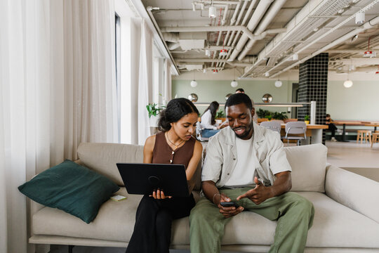 Male worker smiling and pointing at phone showing to female worker holding laptop while they sit on couch
