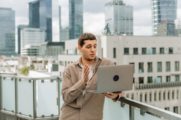 Fototapeta premium Male worker talking and gesturing in front of laptop he holds while standing and leaning on fence