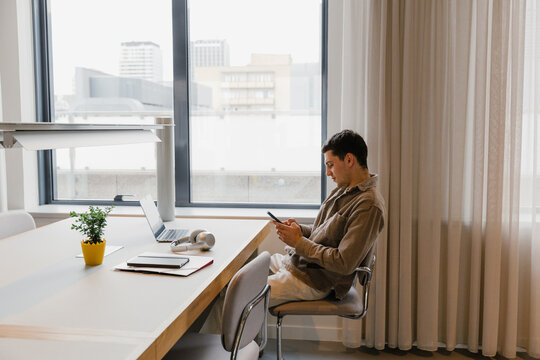 Male worker typing on phone and sitting on chair at desk in front of laptop