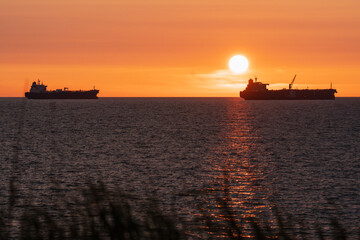 Fototapeta premium View from mainland Estonia of two oil tankers silhouetted against a vibrant golden sunset on the Baltic Sea horizon.