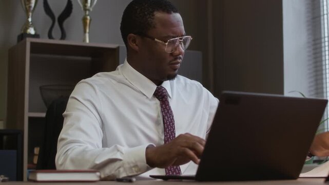 Medium shot of focused young male black principal doing paperwork sitting in front of laptop at desk in school office