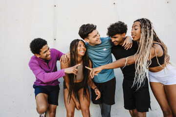 A group of five athletes stand and hug each other while laughing and leaning on a wall while two of them point at each other