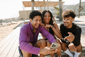 A group of three athletes are talking and laughing while sitting on the floor and looking at a phone that one of them is showing