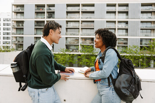 Male and female students talking and holding notebooks while standing and leaning on fence