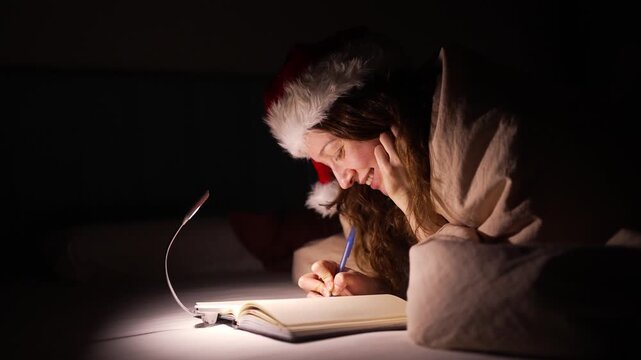 Cheerful young woman in a santa hat lying in bed under the covers at night, smiling while writing a christmas wish list or a letter to santa claus in her notebook with a small lamp