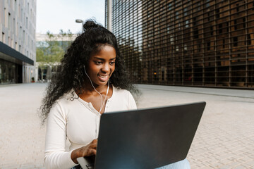Female student typing on laptop and listening to earphones while sitting and smiling