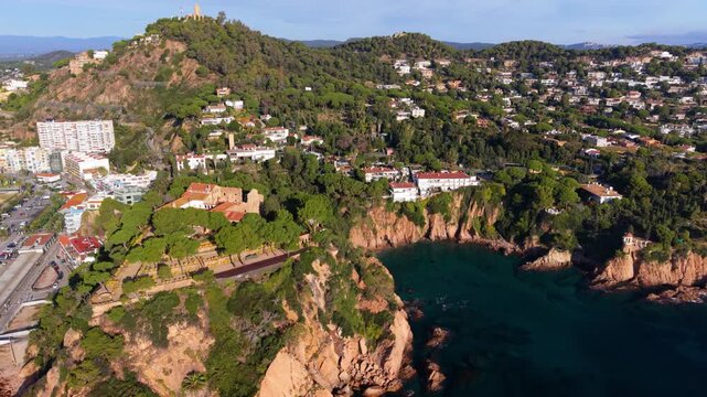 Aerial footage showing the beautiful spanish coastal town of blanes on the costa brava, featuring rocky cliffs, green hills, historic buildings, and the turquoise mediterranean sea