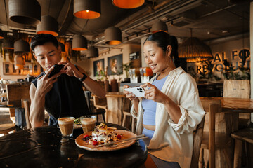 A man and a woman are taking pictures of food and drinks on their phones while sitting at a table and smiling