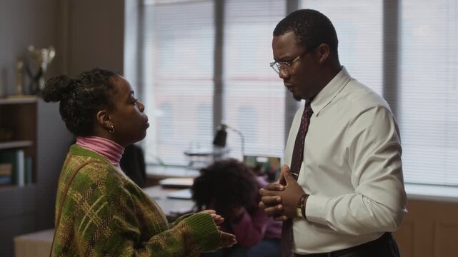 Medium shot of young African American male principal talking to mother of female teenager with misbehavior in school office