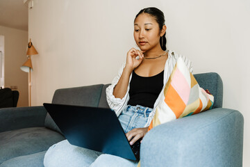 A woman sits on a couch and scrolls through her laptop while looking at it