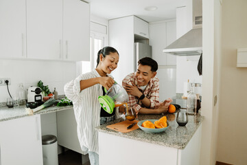 Woman pouring juice from juicer into glass while man holding towel next to her as they stand at table and smile