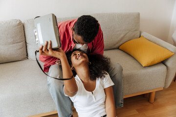 A woman holds a camera and sits on the floor while smiling and looking at a man sitting behind her on the couch