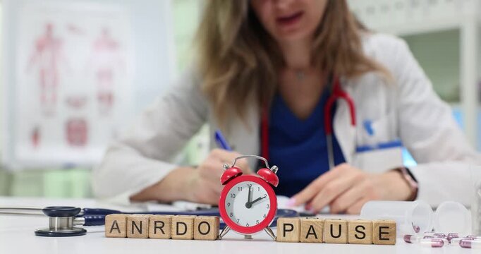 Wooden blocks spell word Andropause near red alarm clock while woman doctor works at desk. Woman specialist writes notes about male hormone problem