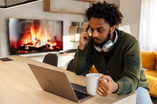 Male musician holding a cup and looking thoughtfully at laptop while sitting at table
