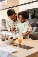 Father smiling and helping his daughter stir with cutlery in a bowl while they stand at the table
