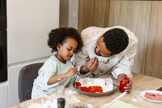 Father holding a jar and talking to his daughter who is sitting at the table and spreading sauce on the dough with a spoon