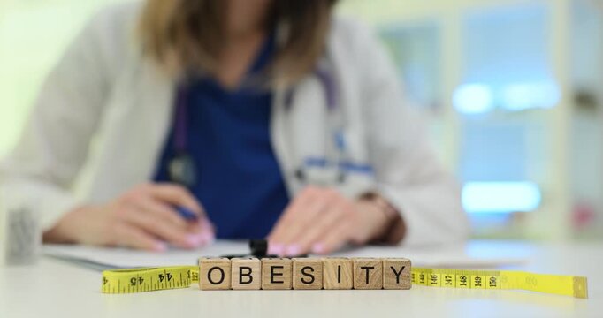 Wooden blocks spell word Obesity near measuring tape and pills while woman doctor writes notes. Woman specialist records data on weight problem