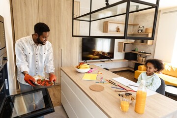 Father standing and showing pizza in baking dish to his daughter sitting at table and they smiling