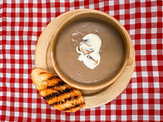 Overhead or flat lay view of a bowl of homemade creamy mushroom soup,garnished with a swirl of cream and sliced mushrooms,with toasted grilled bread on a rustic plate and red checkered tablecloth