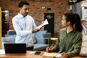 A male employee stands and reads a document while a female employee sitting at a desk listens to him
