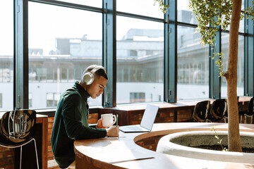 Male worker writing with pencil and holding cup while listening to headphones and sitting at desk in front of laptop