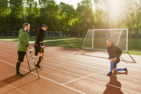 A man stands on one knee and talks while two men stand next to him, leaning on crutches