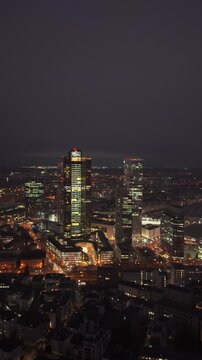 Tripod aerial perspective of Frankfurt's bustling financial district at night in vertical format. Glowing skyscrapers like Tower 185 and city streets create urban scene