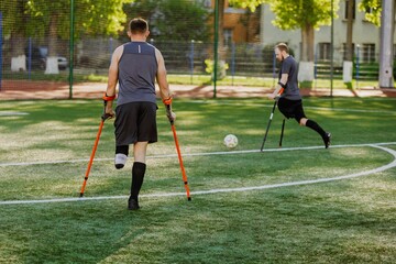 A man walks and holds crutches while looking at a man who is swinging his leg to hit a ball while leaning on crutches
