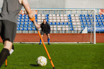 A male goalkeeper stands in the goal and looks at a male player on crutches who is about to kick the ball