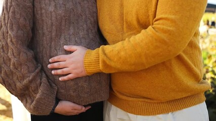 Close-up shot of a pregnant couple standing together outdoors on a sunny autumn day. Man gently places his hand on the woman baby bump as they hug and enjoy warm seasonal light. Concept of family