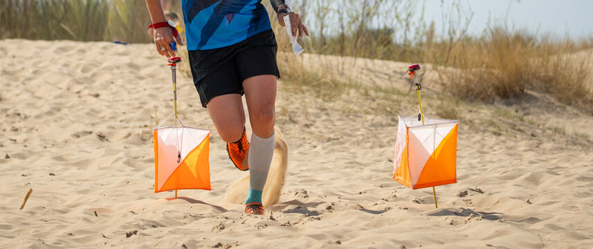 Orienteering athlete running in sandy terrain with two control markers. Close-up action shot of runner's legs on beach dunes. - Powered by Adobe