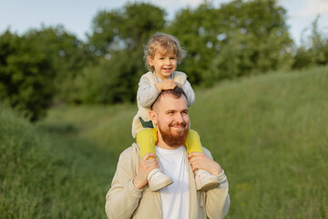 Smiling father with a red beard carrying his toddler on his shoulders while walking outdoors. Warm natural light, joyful family moment and peaceful green landscape.