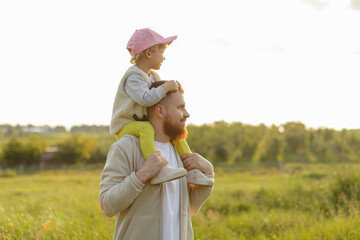 Father carrying toddler on his shoulders while walking outdoors at sunset. Warm natural light, happy family moment and peaceful countryside atmosphere.